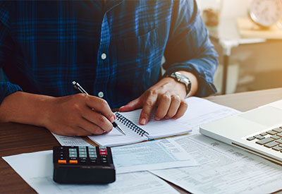 A person in a blue plaid shirt sitting at a desk, writing in a notebook with a pen, surrounded by papers and a calculator, with a laptop partially visible in the background.