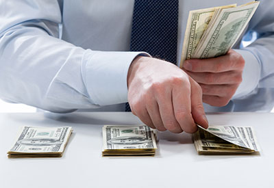 A person wearing a light blue shirt and a dark tie is organizing multiple stacks of U.S. dollar bills on a white surface, holding a fan of bills in one hand while adding to one of the piles—meticulously managing an inheritance that could easily tempt any spendthrift.