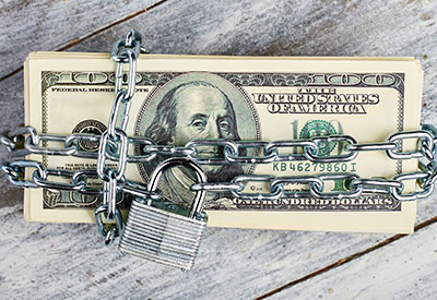 A stack of 100-dollar bills wrapped in metal chains secured with a padlock, symbolizing financial security or restricted access to money. The bundle, potentially an inheritance, is placed on a rustic wooden surface.