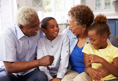 Two elders and two children are sitting on a couch in a living room, smiling and looking at each other. The elders, a man and a woman in casual clothes, discuss the concept of per capita distribution of assets. The children, a boy in a white shirt and a girl in yellow, appear happy and engaged.