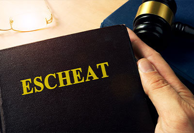 A hand holds a book with the title 'ESCHEAT' in gold letters on the cover. Nearby, there is a gavel resting on a blue book, and a pair of glasses is placed on the table, suggesting an estate planning or legal context.