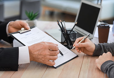 A person in a suit holds a clipboard with a document, pointing at a specific section, while another person, holding a pen, appears ready to sign. The scene, set at a desk with a laptop, coffee cup, and pencils in the background, suggests an important moment in estate planning or inheritance decisions.