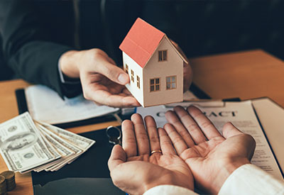 A person hands over a small model house to another, symbolizing a real estate transaction.