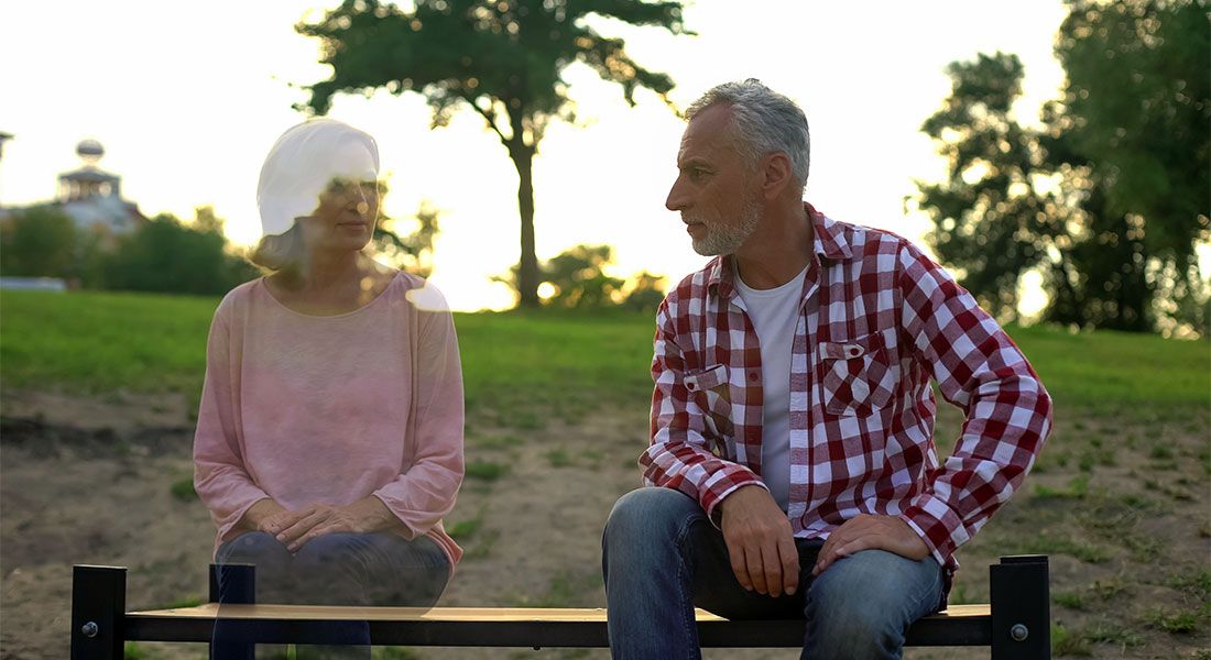 Senior man sitting on a bench looking to his side at the ghost of his wife