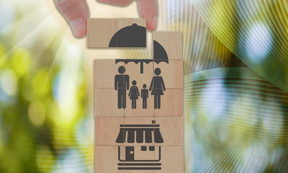 A hand places a block with an umbrella over a family and a business, symbolizing life insurance as a versatile planning tool for families and businesses.
