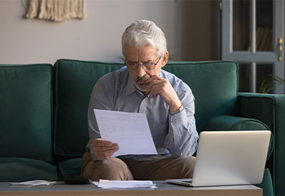 An elderly man with glasses sits on a green sofa, looking at a document in his hands. He appears thoughtful, with one hand touching his chin. A laptop is open on the coffee table in front of him, alongside papers related to estate planning. The setting is a cozy living room.