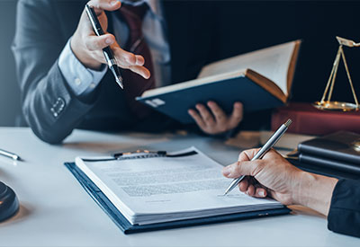 A close-up of two people working at a desk. One person is holding a pen and a book while gesturing, and the other is signing a will. Legal scales and a briefcase are visible in the background, suggesting an estate planning or professional legal setting.