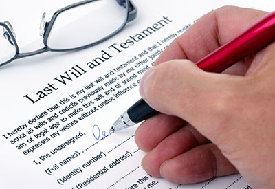 A hand holding a pen is signing a 'Last Will and Testament' document. Glasses are placed beside the document, partially visible. The text on the document includes spaces for personal details such as identity number and residential address.