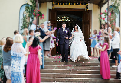 A newlywed couple dressed in formal attire walks down the steps of a building adorned with flowers. Surrounded by cheering friends and family tossing flower petals, the bride holds her dress, smiling, while the groom accompanies her, equally happy—a moment they will cherish forever.