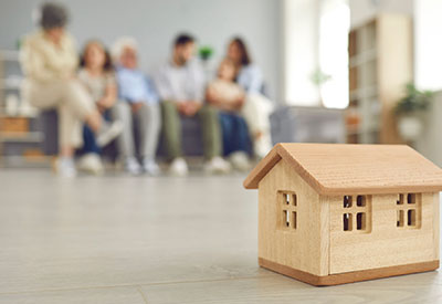 A wooden house model with a blurred family in the background, representing the legacy and asset protection aspects of estate planning for real estate investors.