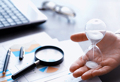 A hand holding an hourglass over financial documents and analysis tools, symbolizing time management in financial planning or legal processes.