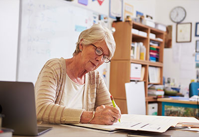 An elderly woman is seated at a desk, writing in a notebook.
