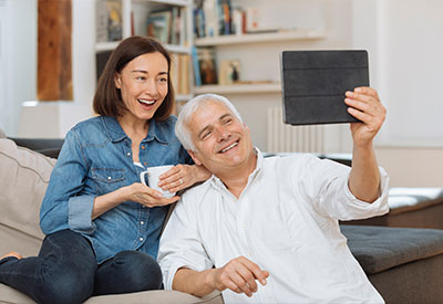 A smiling older couple sits on a couch, using a tablet for a video call.