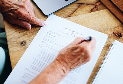 A person's hand is seen signing a last will and testament document on a wooden table, demonstrating careful estate planning.
Another hand is pointing at a specific section of the document, facilitating clear communication. A laptop and notepad are also partially visible in the scene.