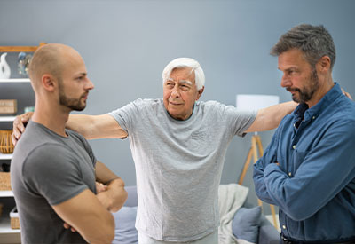 A senior man in a gray shirt stands with his arms outstretched, appearing to mediate an estate planning discussion between two younger men who are facing each other with crossed arms in a modern living room.
