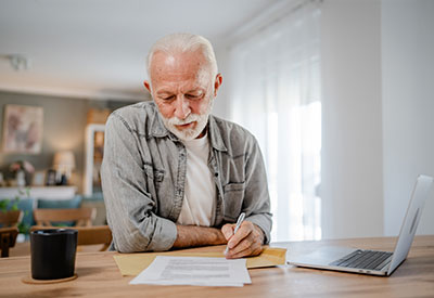An elderly man with a white beard is sitting at a table, focused on writing an estate planning document. He is wearing a denim shirt and a white t-shirt. A laptop and a black mug are on the table in the brightly lit room.
