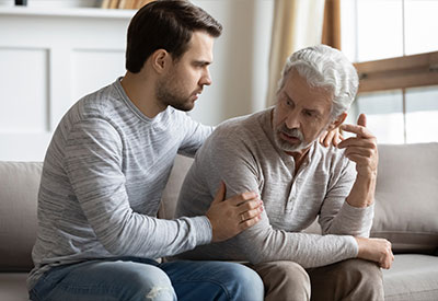A young man conversing and comforting an elderly man on a sofa, both showing serious expressions and emotional engagement in a home setting.