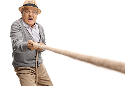 An elderly person in a straw hat and gray sweater appears to be pulling hard on something while expressing surprise, highlighting the struggle for autonomy and control.