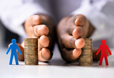 A person in a white shirt uses their hands to support two stacks of coins, with a blue figure beside one stack representing taxes and a red figure beside the other symbolizing investment.