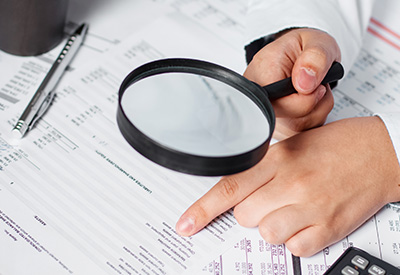 Person examining financial documents related to taxes with a magnifying glass and a calculator nearby.