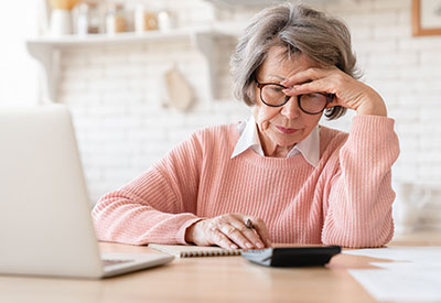An elderly woman, looking stressed while calculating finances, likely considering Medicaid eligibility and asset protection trust options.