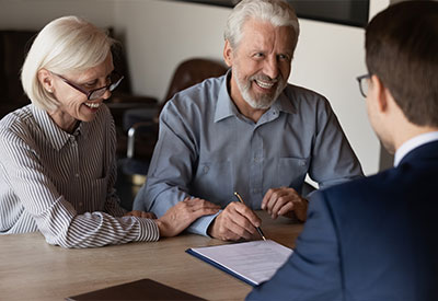 An elderly couple is seated at a desk, smiling and discussing estate planning with a man in a suit who has his back to the camera. The couple appears happy and engaged, with the man holding a pen and looking at a document on the table.