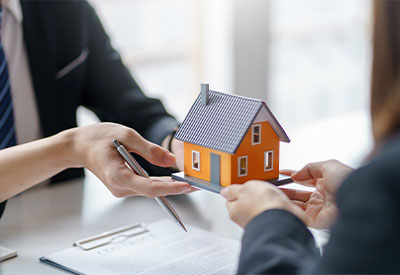 Two people in business attire are seen from the waist up, exchanging a small house model over a desk with papers and a pen. The image likely represents an estate planning or property transaction discussion related to home ownership.