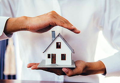 A person wearing a white shirt holds a small model of a house on their left hand while their right hand hovers protectively over it, symbolizing home ownership and security for a home.