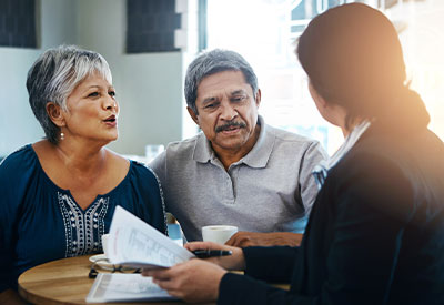 An elderly couple sits at a table in conversation with a business professional. The woman is speaking, and the man is attentively listening. The business professional, holding documents and a pen, discusses estate planning and what measures to take in case of incapacity.