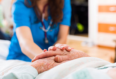 A nurse in blue scrubs gently holds the hand of an elderly patient lying in bed, her stethoscope visible around her neck. The scene conveys a sense of care and support, highlighting the importance of compassion, especially when dealing with issues like incapacity in a medical or caregiving setting.