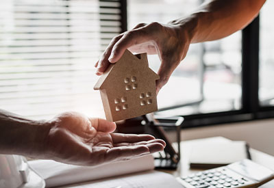 Close-up of two hands exchanging a wooden house model. One hand, extending from the right side of the image, is passing the house to another hand below it, symbolizing real estate transaction or ownership transfer. An office setting is visible in the blurred background, highlighting estate planning nuances.