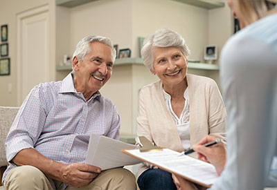 An elderly couple sits on a couch, smiling and holding documents about their revocable trust, while conversing with a person holding a clipboard
