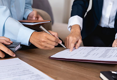 Three people in business attire are gathered around a table, discussing documents. One person is pointing at a document with a pen while another holds a notebook.
