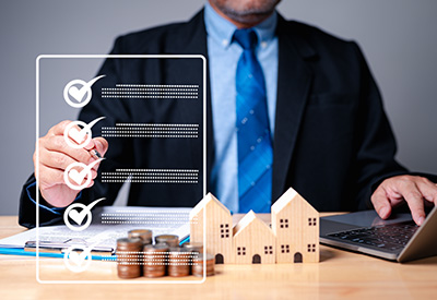 A businessman sitting at a desk with checklist, miniature houses, and coins, symbolizing estate planning components for managing assets and succession.