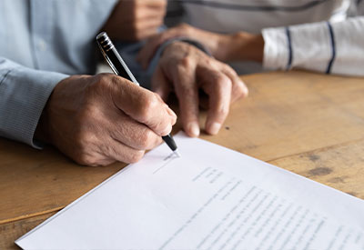 A close-up of an elderly person signing a document related to long-term care costs at a wooden table. Another person, wearing a striped sleeve, gently supports the elder's arm