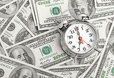 A silver alarm clock rests on a pile of hundred-dollar bills, showing 10:10.