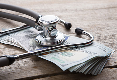 A stethoscope rests on a stack of US hundred-dollar bills alongside documents detailing Medicaid's impact on long-term care costs, all set on a wooden surface.