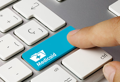 A person presses a blue 'Medicaid' key on a keyboard, symbolizing Medicaid eligibility, asset protection, and estate recovery concerns.