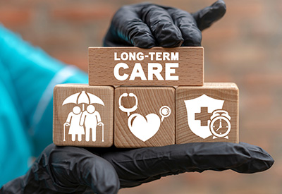 A gloved hand holds wooden blocks labeled 'Long-Term Care' with icons for healthcare and protection