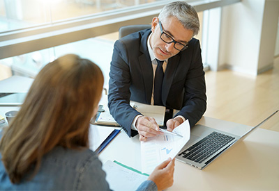 A man in a suit and glasses sits at a desk in a modern office, discussing a document with a woman in a blue jacket. A laptop is open on the desk