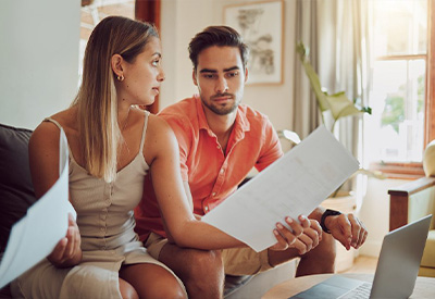 A couple sits on a couch, reviewing documents. The woman holds a paper and looks at the man, who is also holding papers. A laptop is open on the table in front of them.