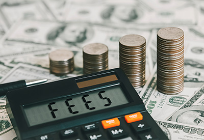 A calculator showing the word 'FEES' on its display is in the foreground. Behind it, stacks of coins increase in height, resting on a background of scattered U.S. dollar bills. A pen lies beside the calculator.