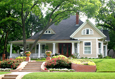 A charming, light beige house with a dark roof and white trim is nestled amidst lush greenery and colorful flowers, an asset to the neighborhood. A brick pathway leads to the porch, framed by two large potted plants, while steps are flanked by vibrant flower beds.