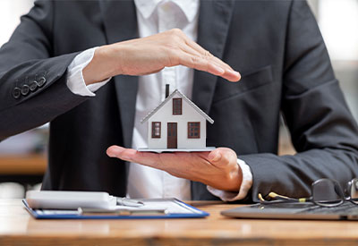 A person in a black suit holds a small white house model, symbolizing the protection of assets. A clipboard with a pen and glasses rests on the wooden table in front of them, embodying the meticulous attention to principal details.