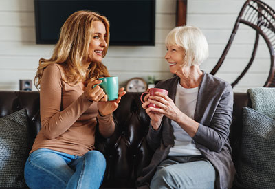 Two women sit on a couch, smiling with mugs in hand. The woman on the left has long blonde hair, wearing a brown top and jeans. The older woman beside her sports short white hair and a gray cardigan over a white shirt.
