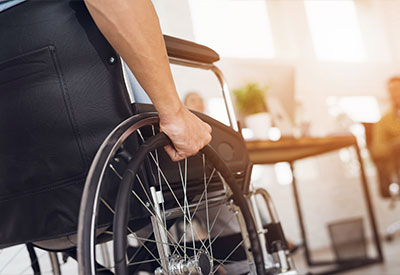 A close-up of a person’s hand on the wheelchair wheel, symbolizing independence. Sunlight streams in, brightening a modern interior where a plant adds life.