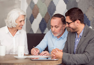 Three people sit at a table discussing estate planning documents. An older woman with white hair smiles, while a middle-aged bearded man writes on paper. A younger bearded man with glasses listens intently. Coffee cups and a tablet are on the table, adding to their focused conversation.