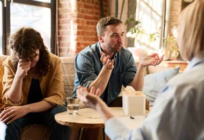 A man speaking animatedly across a table in a cafe, while a woman beside him looks upset