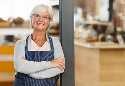 An elderly woman wearing a striped apron, smiles confidently while standing with her arms crossed in a cozy café environment.