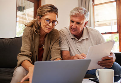 A mature couple reviews estate planning documents together, with the woman using a laptop and the man holding a paper